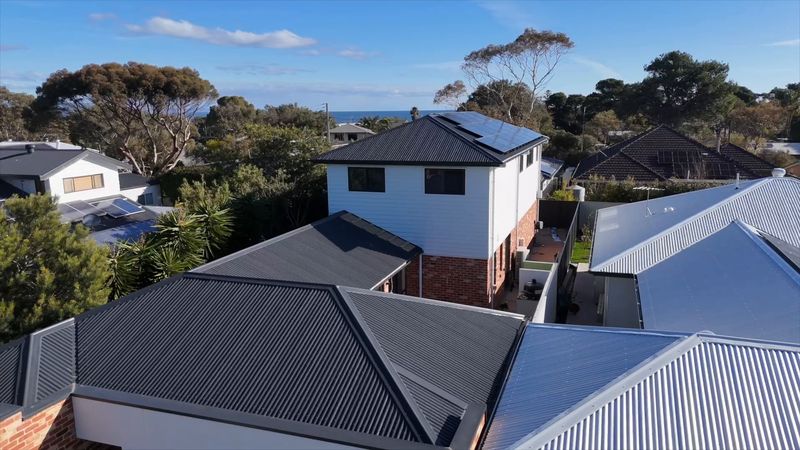 Aerial view of the two-storey home: white weatherboard upper level over red brick, solar on the roof and the sea on the horizon