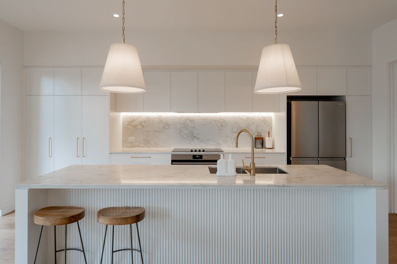 Clapham kitchen front-on: white fluted island bench with timber stools, pendant lights and a lit Eleganto Gold stone splashback